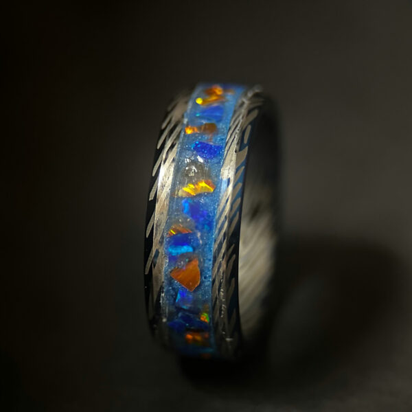 A blue tungsten ring with Damascus pattern and an inlay with blue and orange opal and blue pigment.