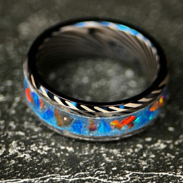 A blue tungsten ring with Damascus pattern and an inlay with blue and orange opal and blue pigment.