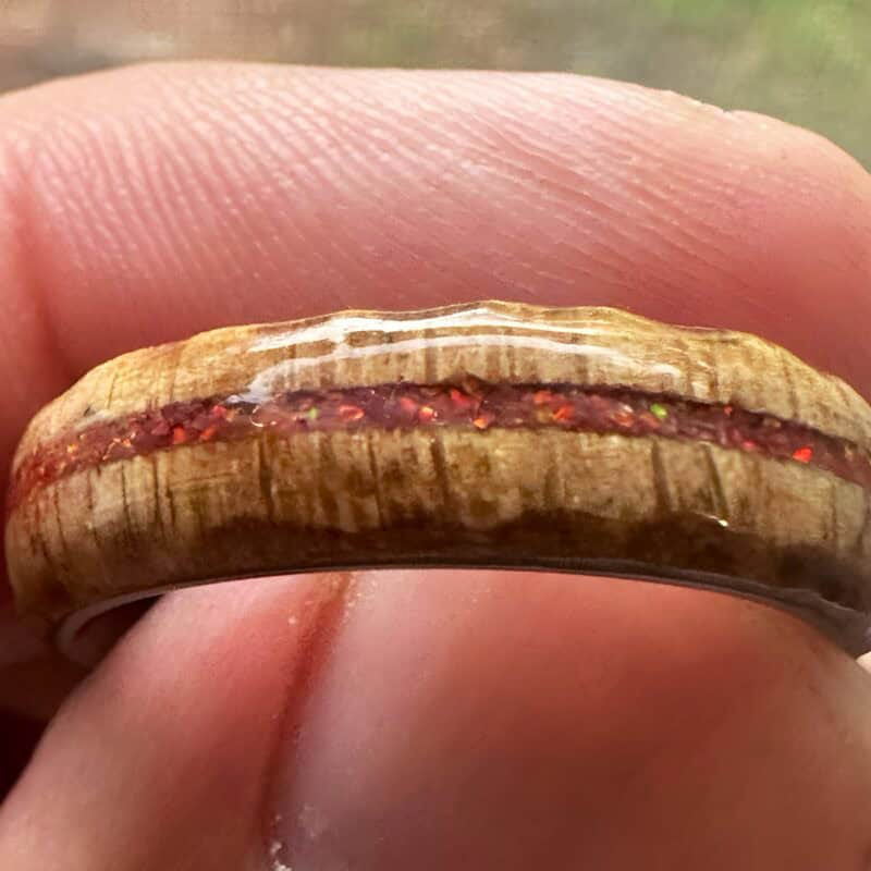 Ambrosia maple ring with red opal inlay, burnt edges, and stainless steel core. Held in fingers