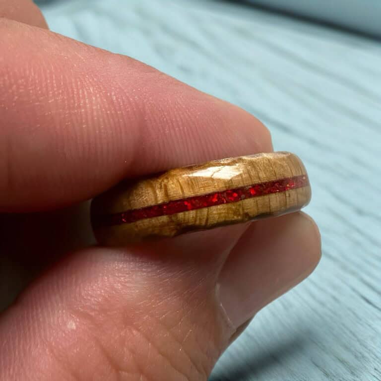 Ambrosia maple ring with red opal inlay, burnt edges, and stainless steel core. Held in fingers over wood board.